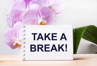 Açık pembe bir orkidenin arka planına karşı masada bir mola al yazısıyla beyaz boş bir defter.