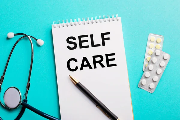 SELF CARE written in a white notepad near a stethoscope, pens and pills on a light blue background. Medical concept