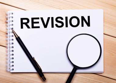 Hafif ahşap bir arka planda bir kalem, bir büyüteç ve REVISION yazılı bir defter yatar.