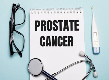 Stetoskop, gözlük ve açık mavi arka planda elektronik termometrenin yanında beyaz bir not defterine yazılmış PROSTATE CANCER. Tıbbi konsept.