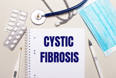 Açık gri arka planda, açık mavi tek kullanımlık yüz maskesi, bir stetoskop, bir elektronik termometre, haplar, bir kalem ve CYSTIC FIBROSIS yazılı bir defter. Tıbbi konsept