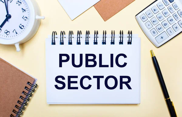 Hafif bir arkaplanda, beyaz bir çalar saat, bir hesap makinesi, bir kalem ve PUBLIC SECTOR yazılı bir defter. Düz yatıyordu