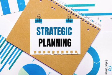 Masaüstünde mavi ve açık mavi grafikler ve diyagramlar, kahverengi bir defter ve mavi ataçlı bir kağıt ve STRATEGIC PLANNING metni bulunmaktadır. Yukarıdan bak. İş konsepti