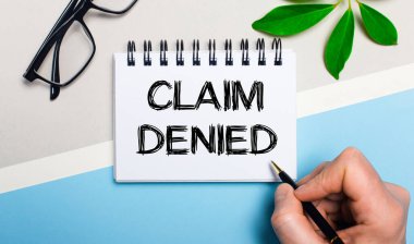 Gri-mavi arka planda, gözlüklerin yanında ve bir bitkinin yeşil yaprağında bir adam bir parça kağıda CLAIM DENIED metnini yazar. Düz yatıyordu. Yukarıdan görüntüle.