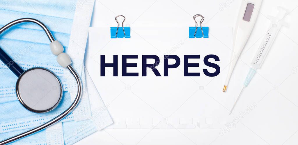 On a light background lie a stethoscope, an electronic thermometer, a syringe, a face mask and a sheet of paper with the text HERPES. Medical concept