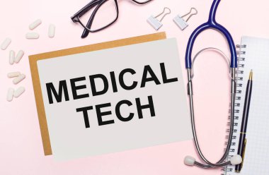 Açık pembe bir arkaplanda, steteskop, beyaz haplar ve kağıt raptiyeler, siyah çerçeveli gözlükler ve MEDICAL TECH yazılı bir kağıt. Yukarıdan bak. Tıbbi konsept
