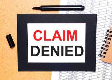 Ahşap bir masanın üzerinde siyah bir keçeli kalem, kahverengi bir not defteri ve siyah çerçeveli CLAIM DENIED yazılı bir kağıt var. Yukarıdan görüntüle.