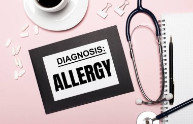 Açık pembe arka planda, kalem, steteskop, beyaz haplar, ataçlar ve içinde ALLERGY yazılı bir kağıt olan bir defter. Tıbbi konsept