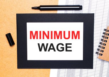 Ahşap bir masanın üzerinde siyah bir açık işaret, kahverengi bir not defteri ve siyah çerçeveli MINIMUM WAGE yazılı bir kağıt var. Yukarıdan görüntüle.