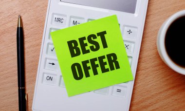 Tahta bir masanın üzerinde beyaz bir fincanda kahve, bir kalem ve üzerinde en iyi teklifin yazılı olduğu yeşil etiketli beyaz bir hesap makinesi var. İş konsepti