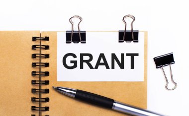Hafif bir arkaplanda, kahverengi bir defter, bir kalem, siyah raptiyeler ve içinde GRANT yazılı bir beyaz kart.
