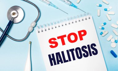 Açık mavi arka planda, elektronik termometre, stetoskop, beyaz ve mavi haplar, şırıngalar ve üzerinde DUR HALITOSIS yazan bir defter. Tıbbi konsept