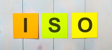 Açık mavi ahşap arka planda ISO yazılı üç renkli etiket.