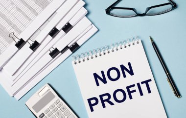 Açık mavi arkaplanda belgeler, gözlük, hesap makinesi, kalem ve NON PROFIT metninin olduğu bir defter var. İş yerinin yakın çekimi. İş konsepti