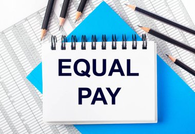 Masanın üzerinde mavi arka planda siyah kalemlerin yanında EQUAL PAY yazılı beyaz defter ve raporlar. İş konsepti