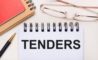 Hafif arkaplanda altın çerçeveli gözlükler, bir kalem, kahverengi ve kırmızı not defterleri ve içinde TENDERS yazan beyaz bir defter var. İş konsepti