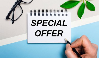 On a gray-blue background, near glasses and a green leaf of a plant, a man writes on a piece of paper the text SPECIAL OFFER. Flat lay. View from above.