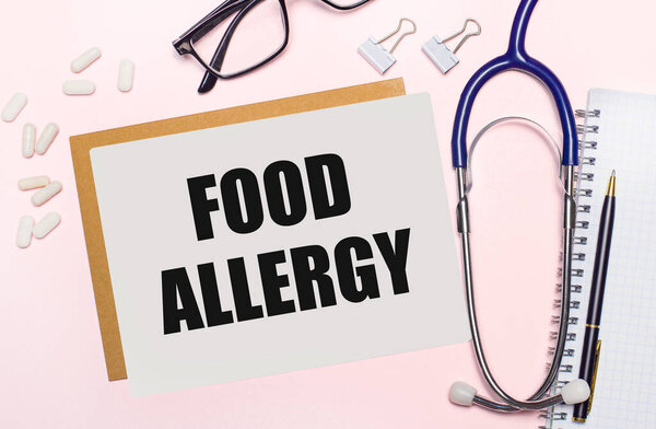 On a light pink background, a notebook with a pen, stethoscope, white pills, paper clips and a sheet of paper with the text FOOD ALLERGY. Medical concept