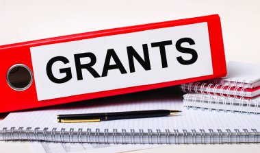 Masaüstünde not defterleri, bir kalem ve içinde GRANTS metni olan kağıtlar için kırmızı bir klasör var. İş konsepti