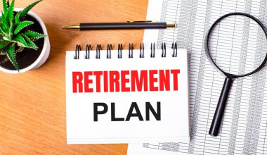 On a wooden table there are reports, a potted plant, a magnifying glass, a black pen and a notebook with the text RETIREMENT PLAN. Business concept