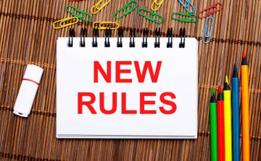 Ahşap bir masada, çok renkli kalemler, ataçlar, beyaz bir flaş bellek ve üzerinde yeni kurallar yazan bir defter. Düz yatıyordu