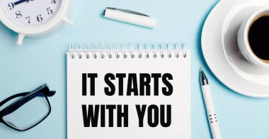 Açık mavi arka planda, kahveli beyaz bir kupa, beyaz bir çalar saat, beyaz bir kalem ve seninle başlayan bir not defteri. Yukarıdan görüntüle