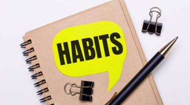 Hafif bir arkaplanda kahverengi bir defter, siyah bir kalem ve ataç ve içinde HABITS yazan sarı bir kart. İş konsepti.