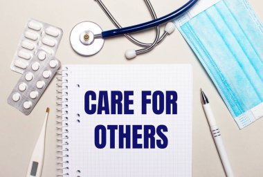 Açık gri bir arka planda, açık mavi tek kullanımlık yüz maskesi, stetoskop, elektronik termometre, haplar, bir kalem ve diğerleri için bir not defteri. Tıbbi konsept