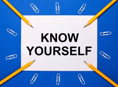 Mavi arka planda, beyaz ataçlar, sarı kalemler ve kendini tanı yazılı beyaz bir kağıt.