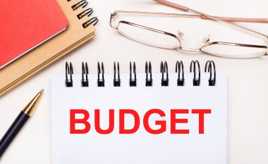 On a light background - glasses in gold frames, a pen, brown and red notepads and a white notebook with the text BUDGET. Business concept