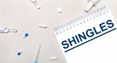On a light background, a syringe, a stethoscope, vials of medicine, an ampoule and a white notepad with the text SHINGLES. Medical concept