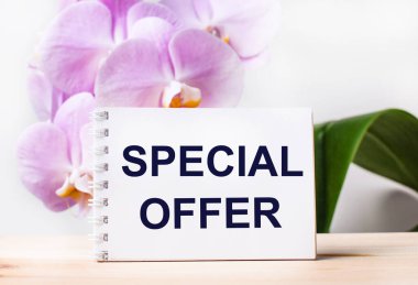 White blank notebook with the text SPECIAL OFFER on the table against the background of a light pink orchid.