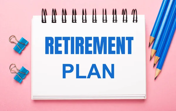 On a light pink background, light blue pencils, paper clips and a white notebook with the text RETIREMENT PLAN