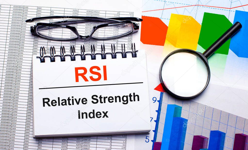 On the desktop are glasses, a magnifying glass, color charts and a white notebook with the text RSI Relative Strength Index. Business concept. View from above
