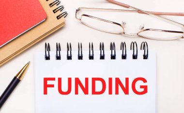 Hafif bir arkaplanda altın çerçeveli gözlük, bir kalem, kahverengi ve kırmızı not defterleri ve üzerinde yazı yazan beyaz bir defter. İş konsepti