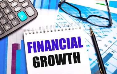 Masaüstü renkli tablolar, bir hesap makinesi, bir gözlük, bir kalem ve FINANCIAL GROWTH metin içeren bir defter içerir
