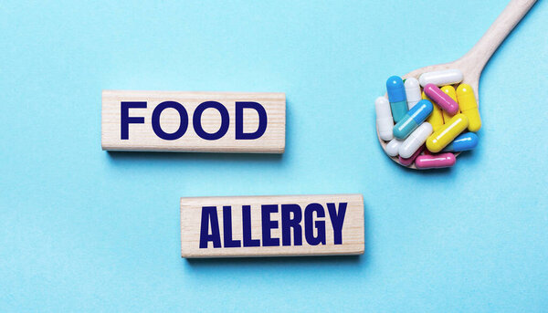 On a light blue background, bright multi-colored pills in a spoon and two wooden blocks with the text FOOD ALLERGY. Medical concept