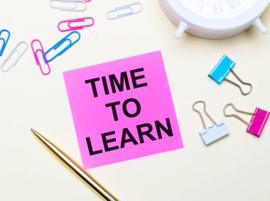 Hafif bir arka planda, beyaz bir çalar saat, pembe, mavi ve beyaz ataçlar, altın bir kalem ve üzerinde Time to EARN yazan pembe bir çıkartma.