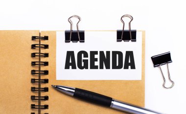 Hafif arkaplanda, kalem, siyah ataç ve AGENDA yazılı beyaz kart içeren kahverengi bir defter.