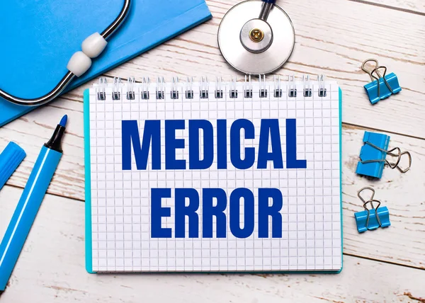 On a light wooden background, a stethoscope, a blue notepad, blue paper clips, a blue marker and a sheet of paper with the text MEDICAL ERROR. Medical concept