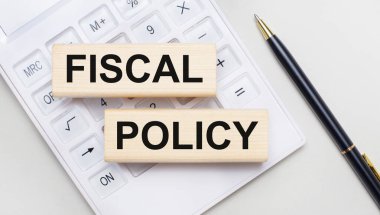FISCAL POLICY metni ile tahta bloklar beyaz bir hesap makinesinde hafif bir arkaplan üzerinde yatmaktadır. Yakınlarda siyah bir kol var. İş konsepti