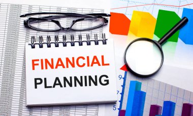 Masaüstünde bir gözlük, bir büyüteç, renk çizelgeleri ve içinde FINANCIAL PLANNING yazan beyaz bir defter var. İş konsepti. Yukarıdan görüntüle