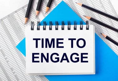 Masanın üzerinde Time to ENGage yazılı beyaz defter mavi arka planda siyah kalemlerin yanında ve raporlar. İş konsepti