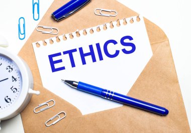 Hafif bir arka planda, bir el işi zarfı, bir çalar saat, ataçlar, mavi bir kalem ve ETHICS yazılı bir kağıt..