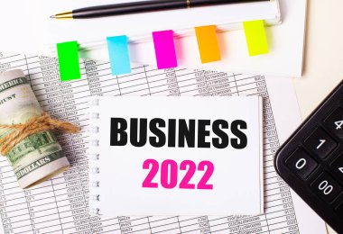 Hafif bir arka planda, bir kalem, dolarlar, içinde BUSINESS 2022 ve parlak çıkartmalar olan bir defter. İş konsepti