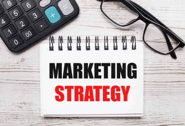 Açık tahta bir hesap makinesi, gözlük ve üzerinde MARKETING STRATEGY yazan boş bir not defteri. İş konsepti