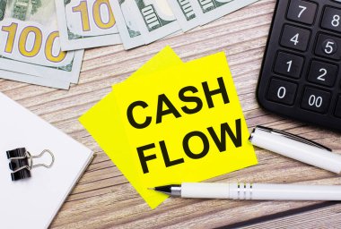 Tahta masada bir hesap makinesi, banknotlar, bir kalem, ataçlar ve CASH FLOW yazılı sarı çıkartmalar var. İş konsepti
