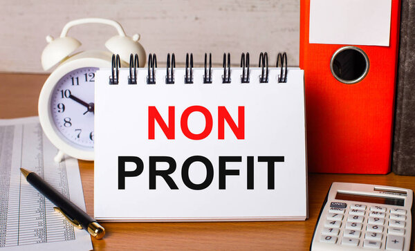 On the table are reports, a white alarm clock, a calculator, folders for papers, a pen and a white notebook with the NON PROFIT. Business concept