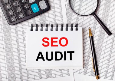 Masada raporlar, bir büyüteç, bir hesap makinesi, bir kalem ve SEO AUDIT yazılı beyaz bir defter var. İş konsepti