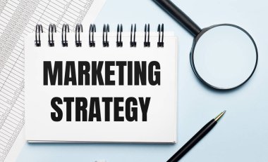 Açık mavi arka planda, raporlar, bir büyüteç, bir kalem ve üzerinde MARKETING STRATEGY yazan bir defter. İş konsepti. Düz yatıyordu.